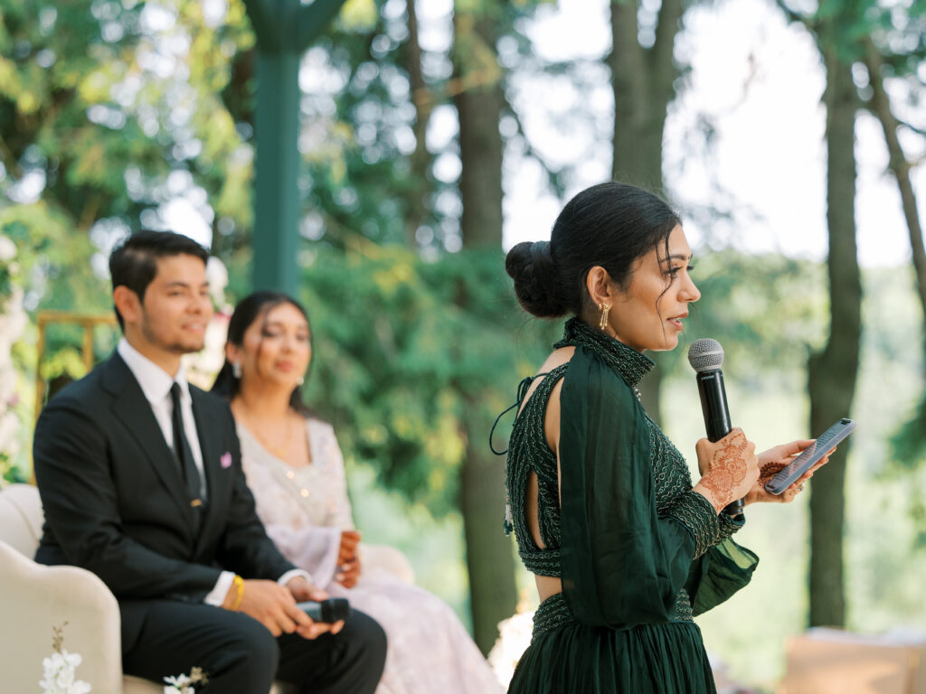 Bride's sister gives sentimental wedding toast while in a green lengha, with bride and groom behind her. 