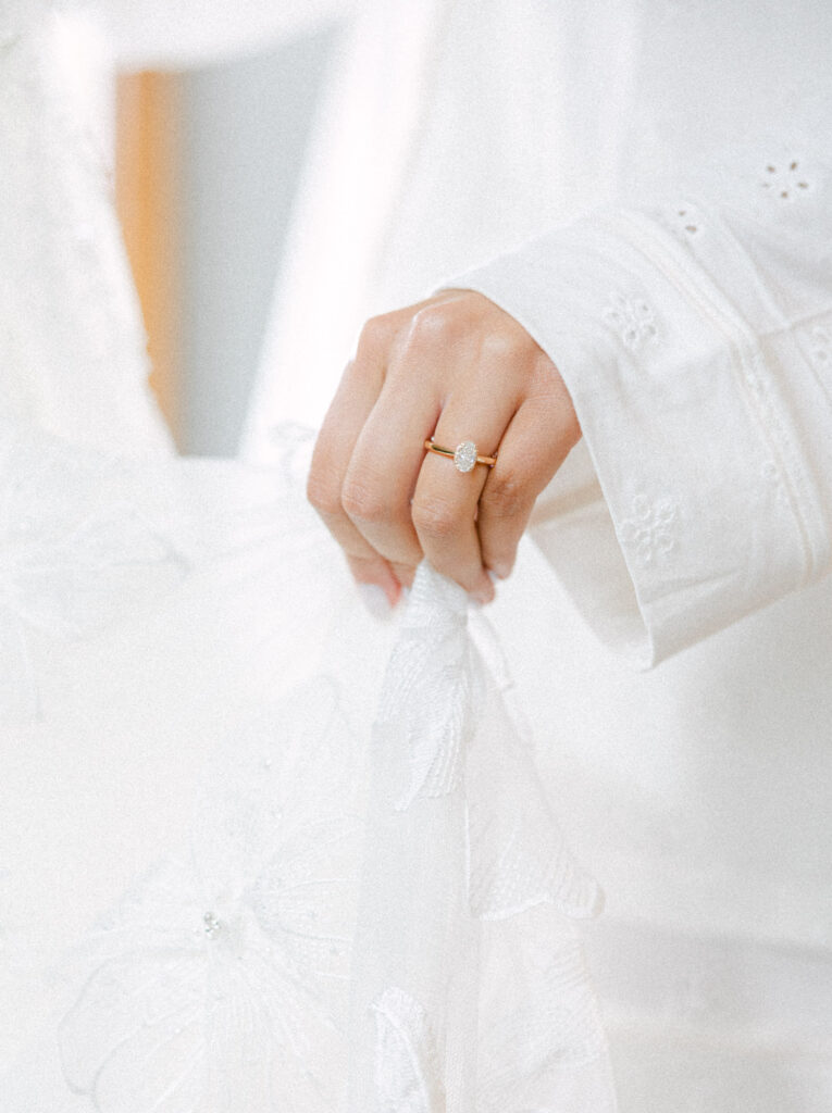 Close-up of a hand wearing a gold ring with an oval stone, gently holding delicate white fabric with embroidered floral patterns. Elegant and serene.