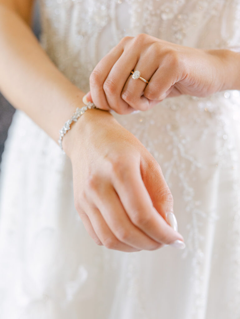Close-up of a hand wearing a gold ring with an oval stone, gently holding delicate white fabric with embroidered floral patterns. Elegant and serene.