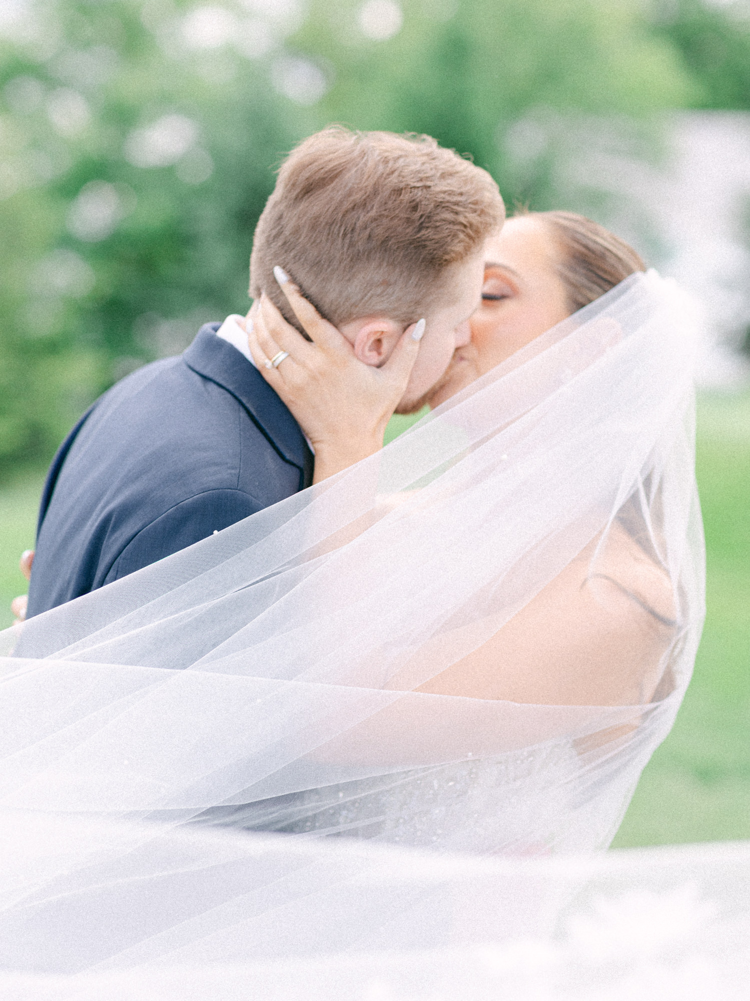 A bride with a long veil and groom in a navy suit share a kiss outdoors, with the bride’s veil draped around them and greenery blurred in the background.